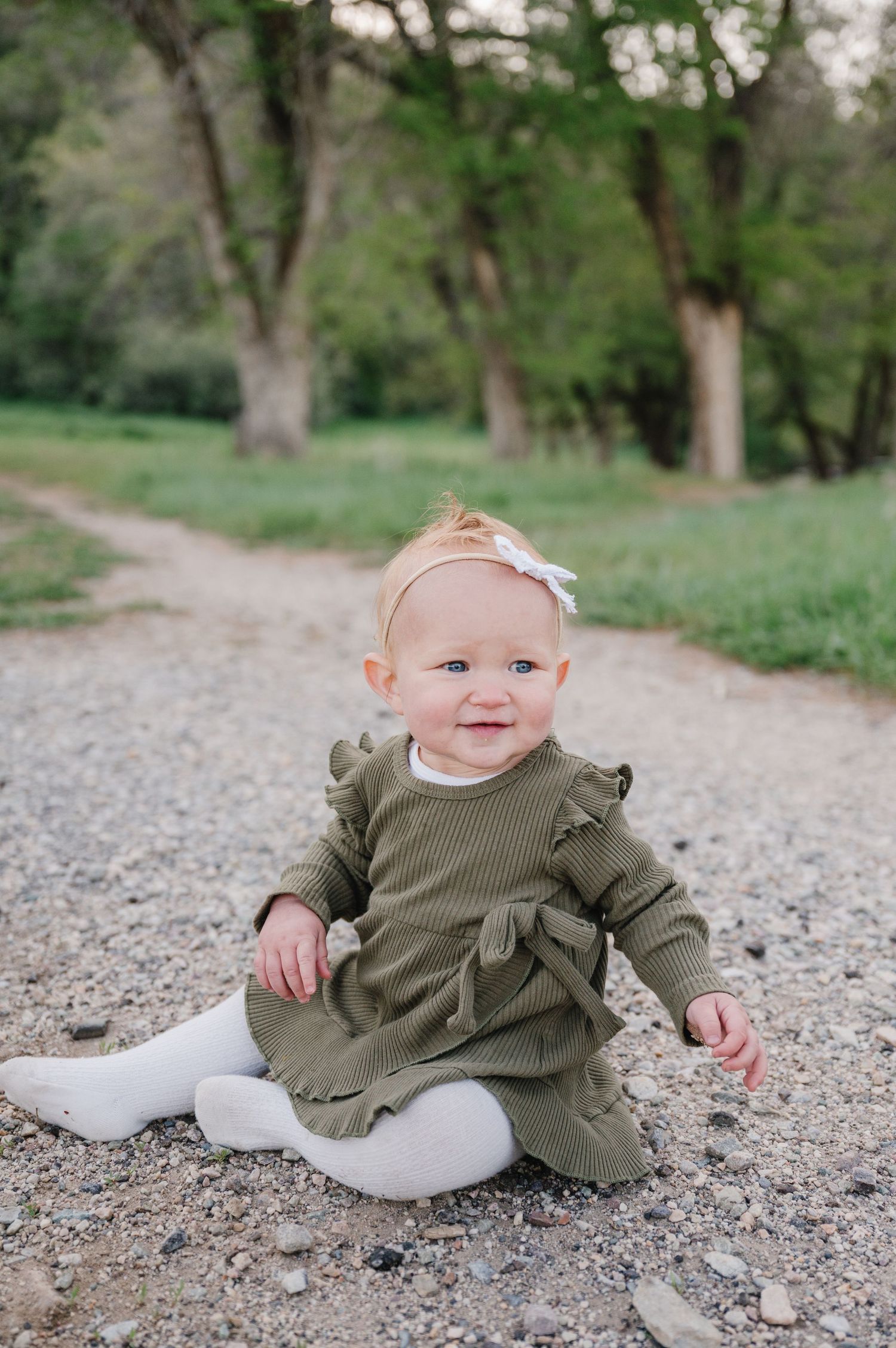 Small child wearing an olive green ruffled dress and white tights sits on a dirt path surrounded by green trees.
