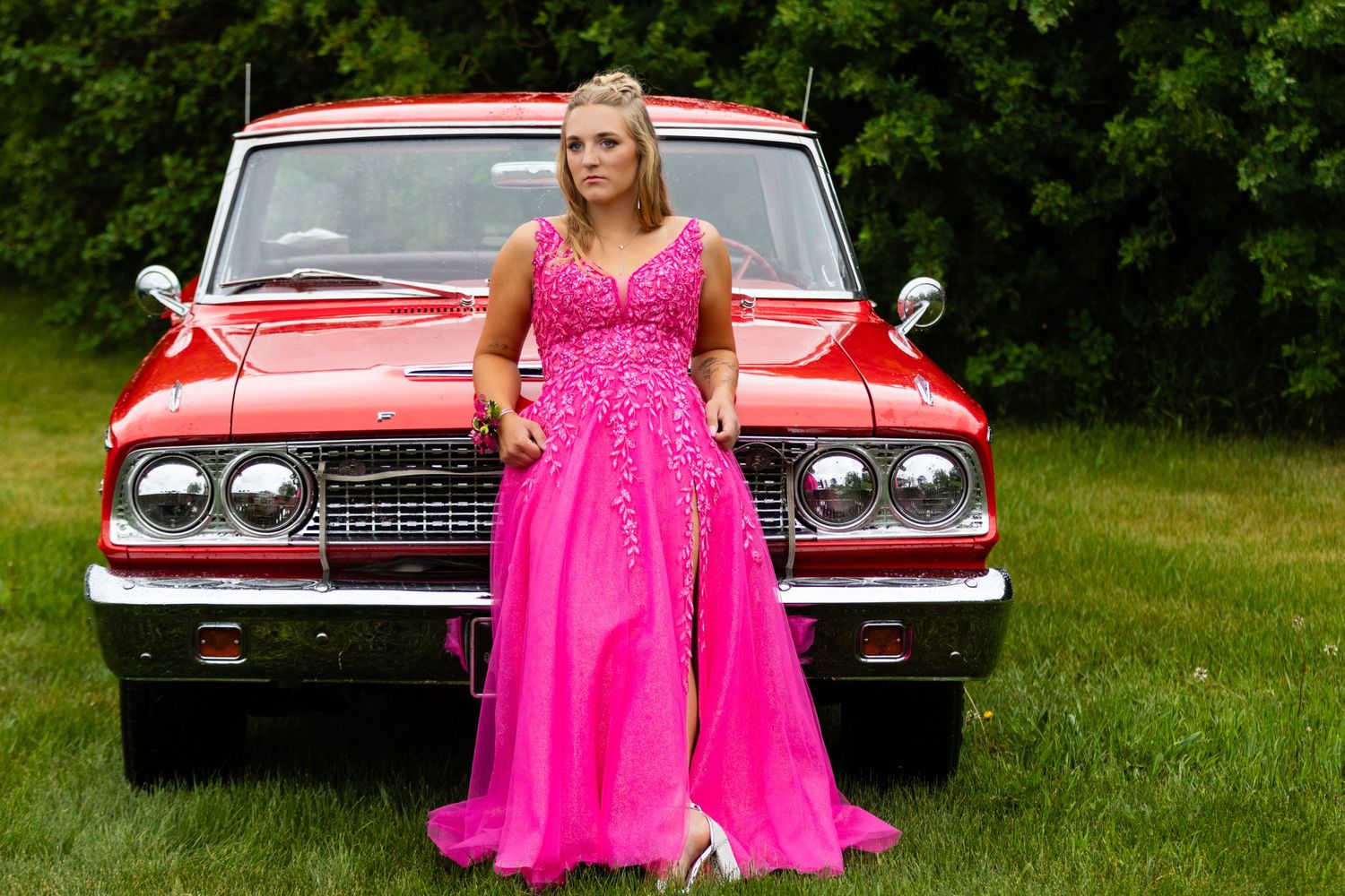 A bright pink formal gown poses next to a classic red Chevrolet car on green grass.