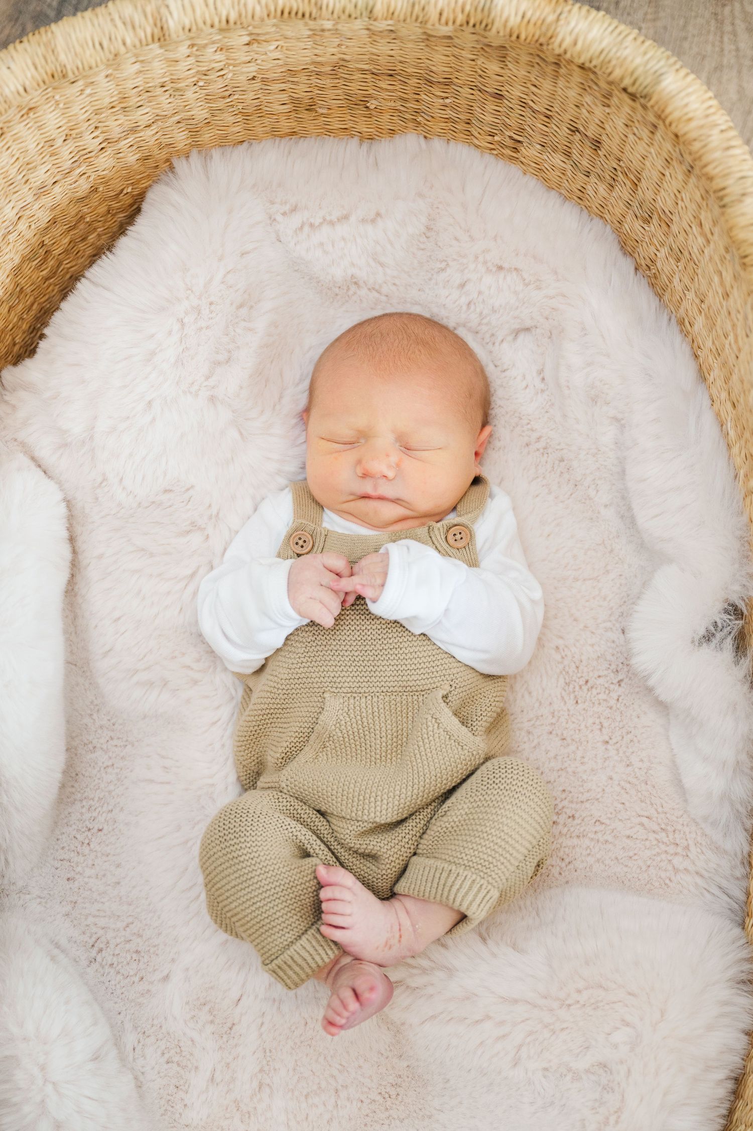 Sleeping newborn baby wearing tan overalls rests in cozy white basket.