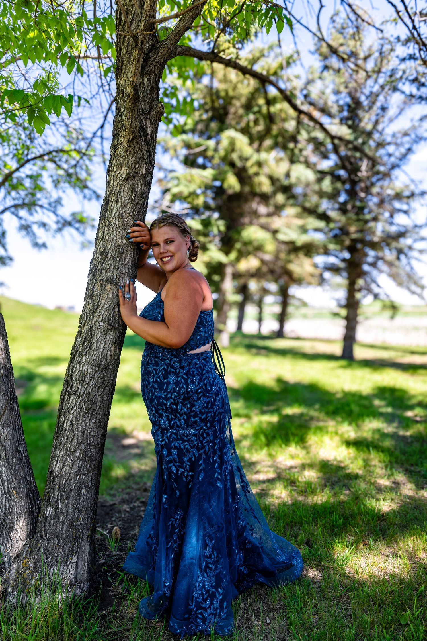 Formal portrait against a tree in a blue lace gown with springtime backdrop.