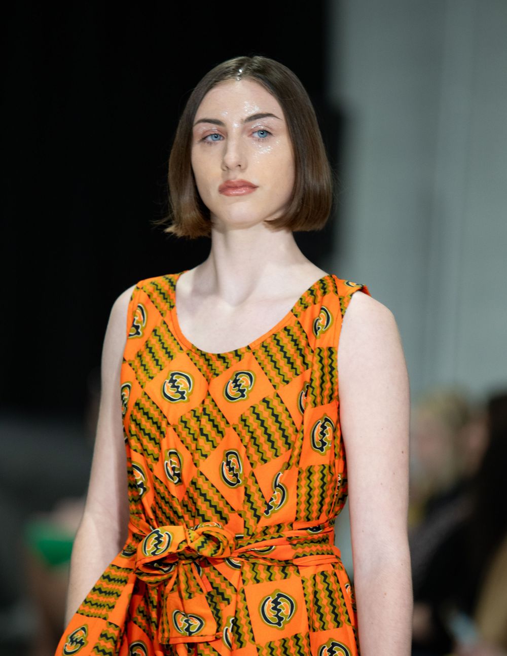 Model showcases orange patterned sleeveless dress with tied waist detail.