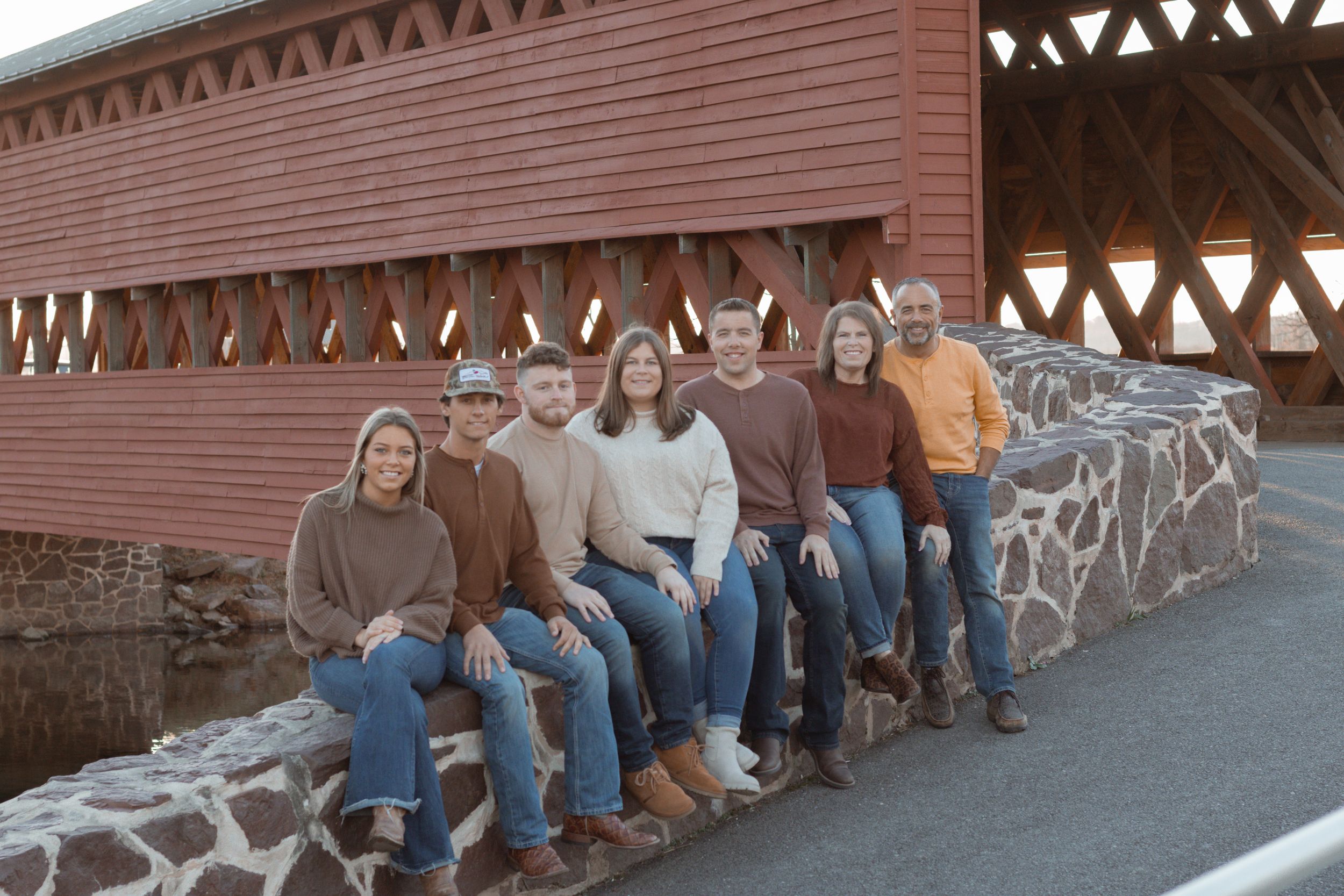A group sitting casually on a stone wall in front of a red covered bridge on an autumn day.
