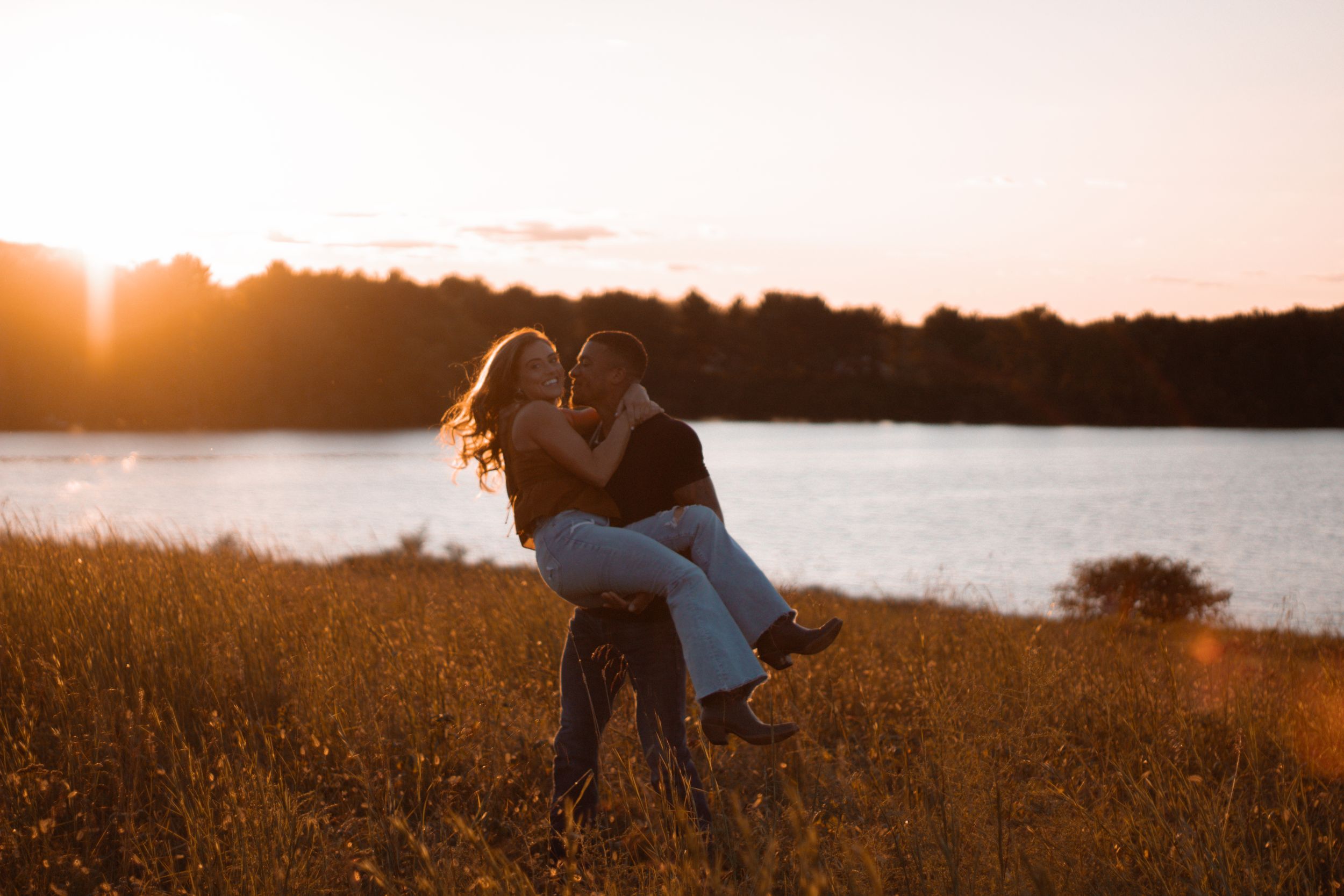 Couple dancing in a sun-lit field beside a lake during golden hour.