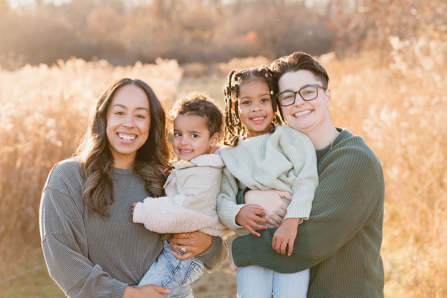 Two adults and two young children pose together smiling in a golden field during a warm autumn family portrait session.