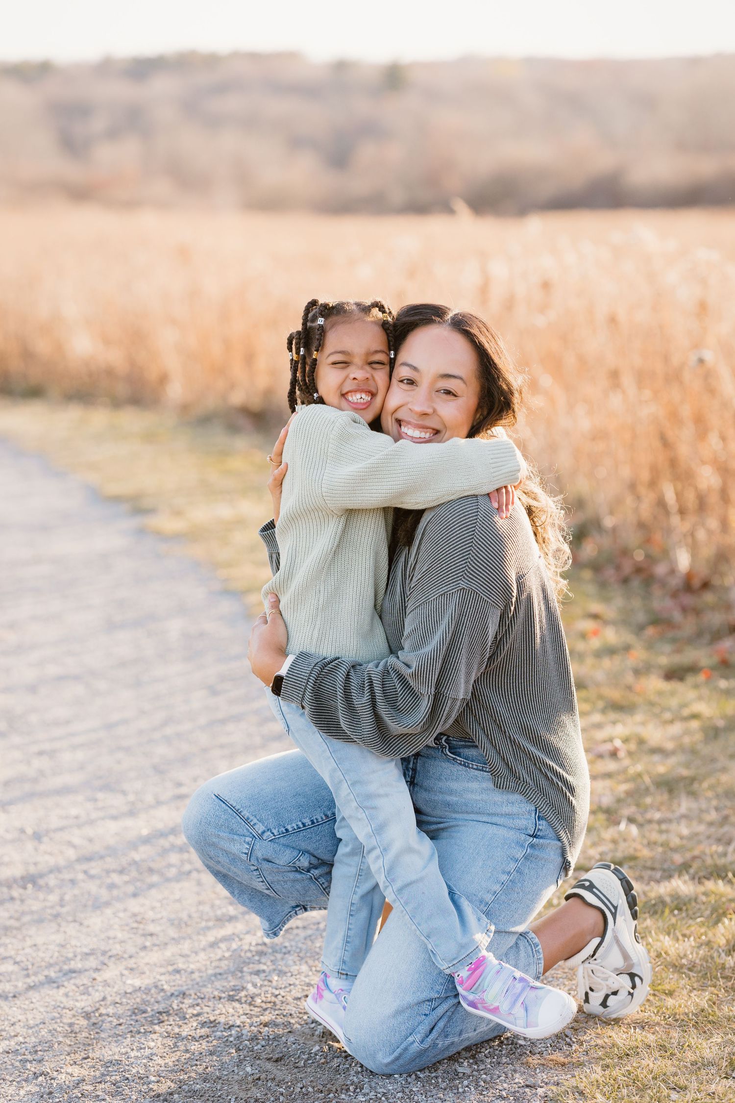 A woman kneels on a path hugging a young girl, both smiling, with golden fields in the background at sunset.