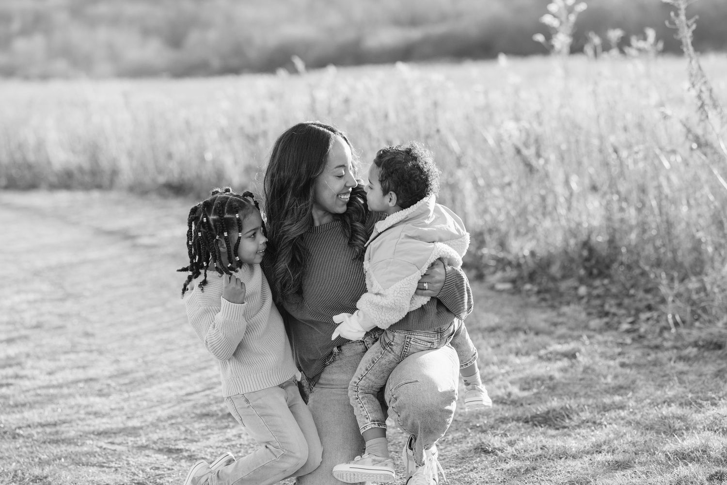 Black and white photo of a mother kneeling in a field, lovingly embracing her two young children outdoors.