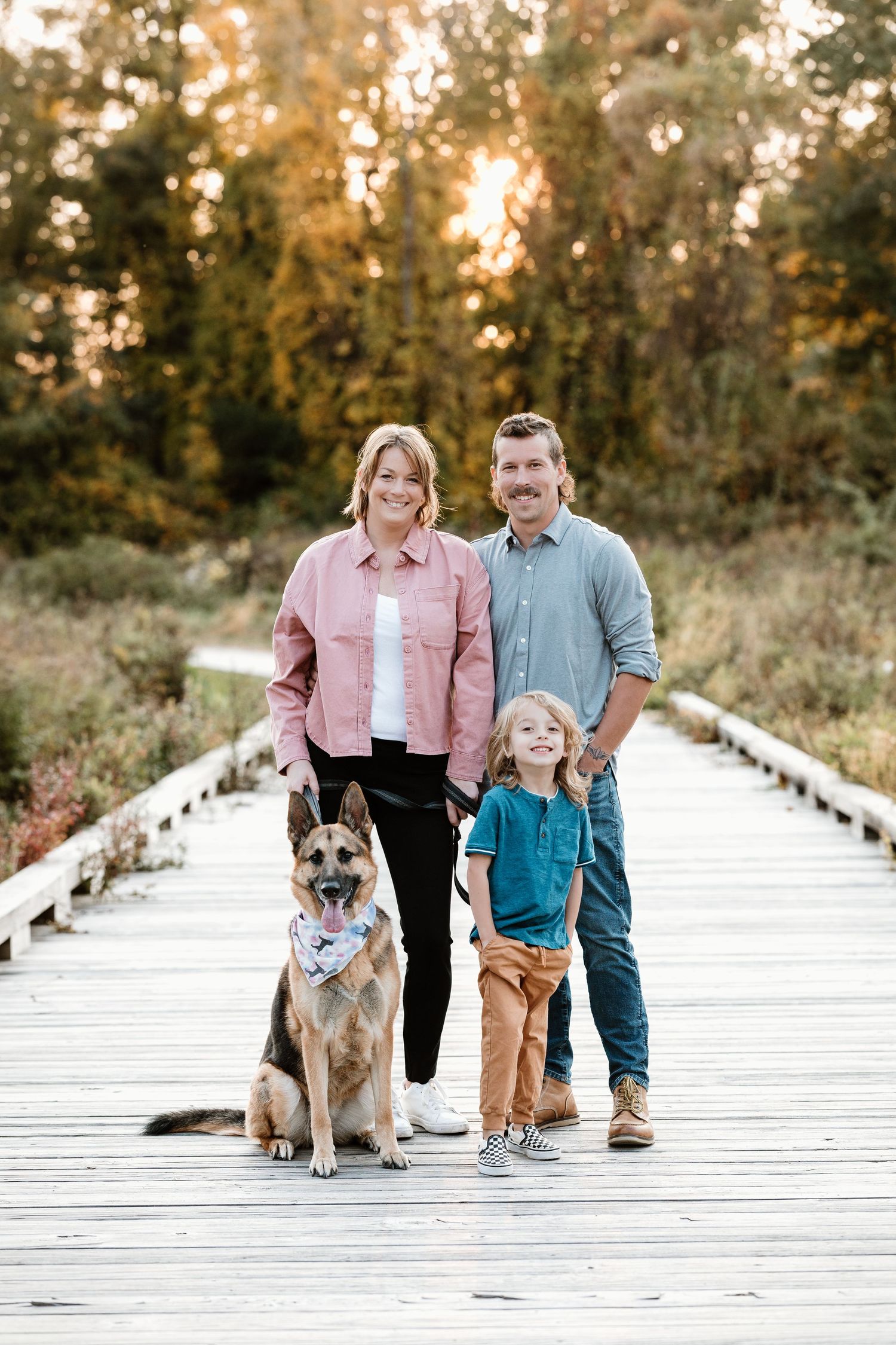 Family and dog pose together on wooden boardwalk during sunset golden hour.