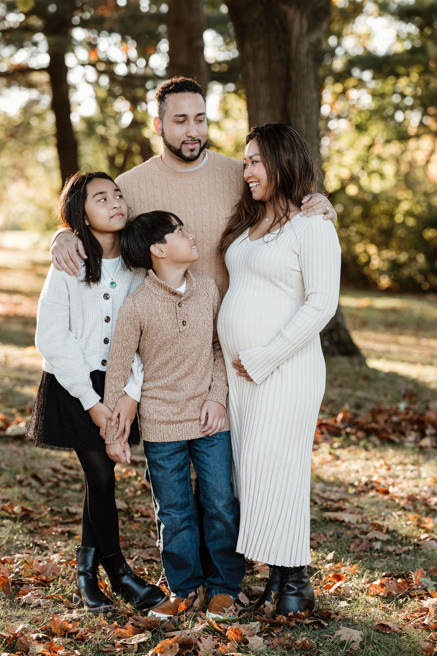 An expectant family poses together outdoors among fall foliage.