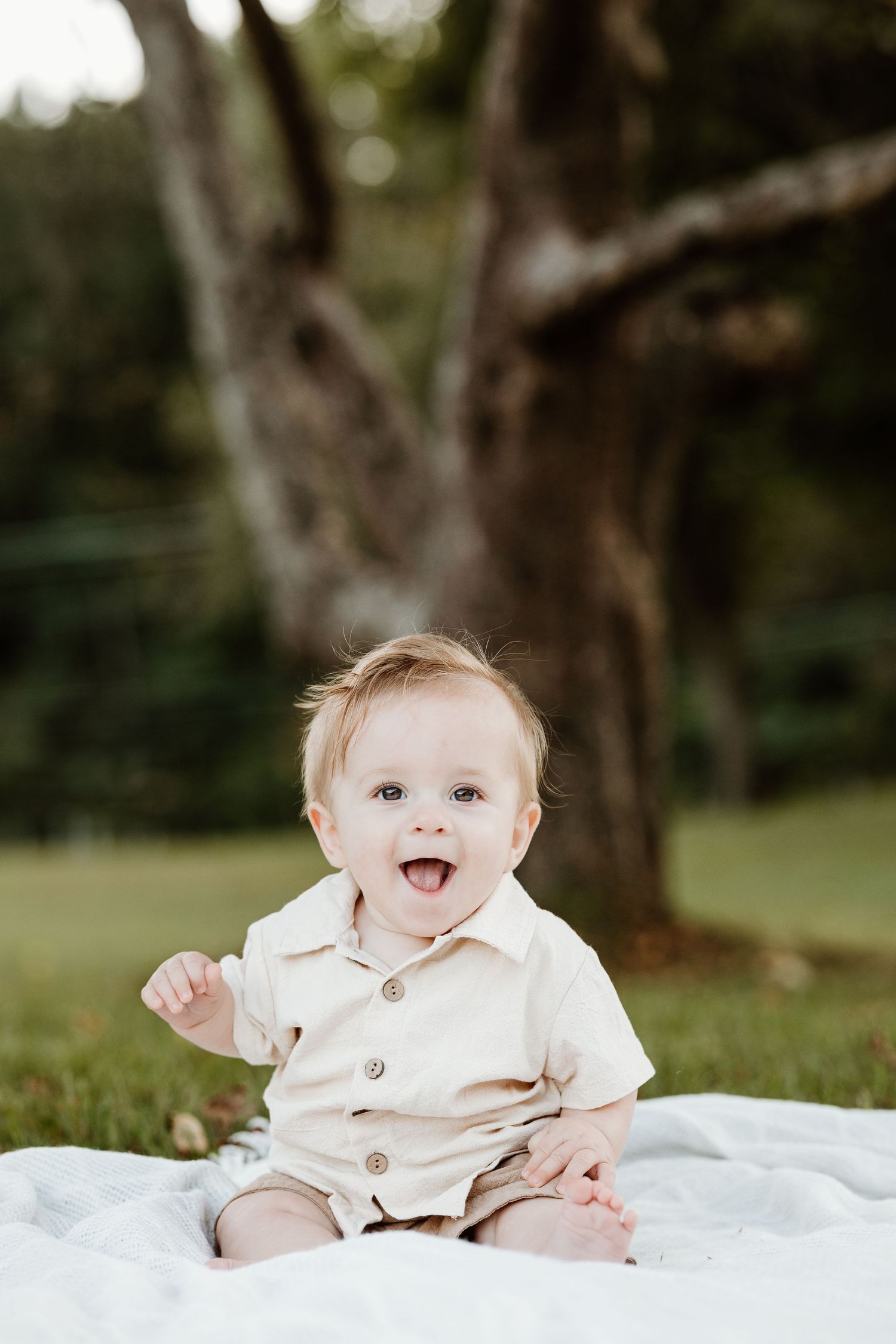 A baby in cream-colored outfit sits on blanket outdoors with joyful expression.