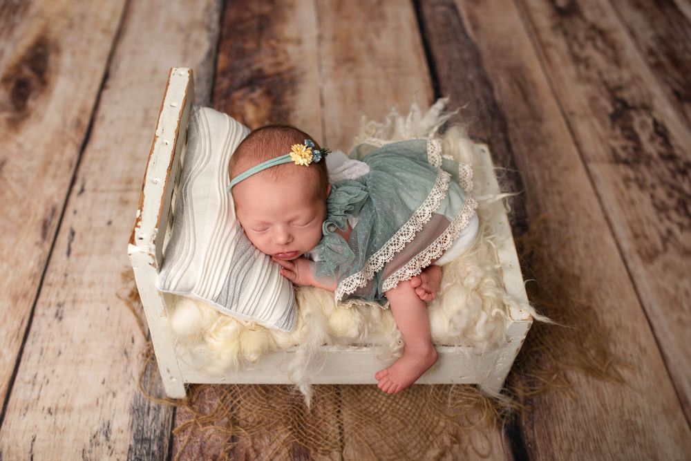 Sleeping newborn in teal outfit on miniature white wooden bed prop.