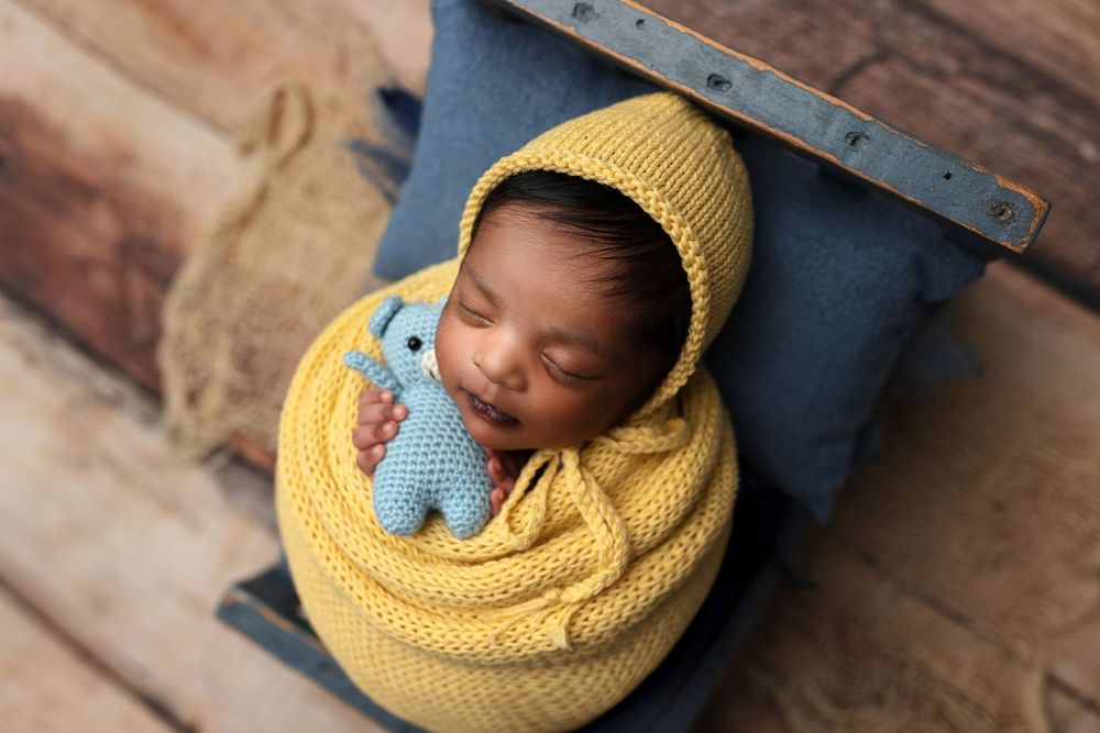 Smiling newborn baby wrapped in yellow knit blanket and bonnet, holding blue stuffed toy in a wooden box.