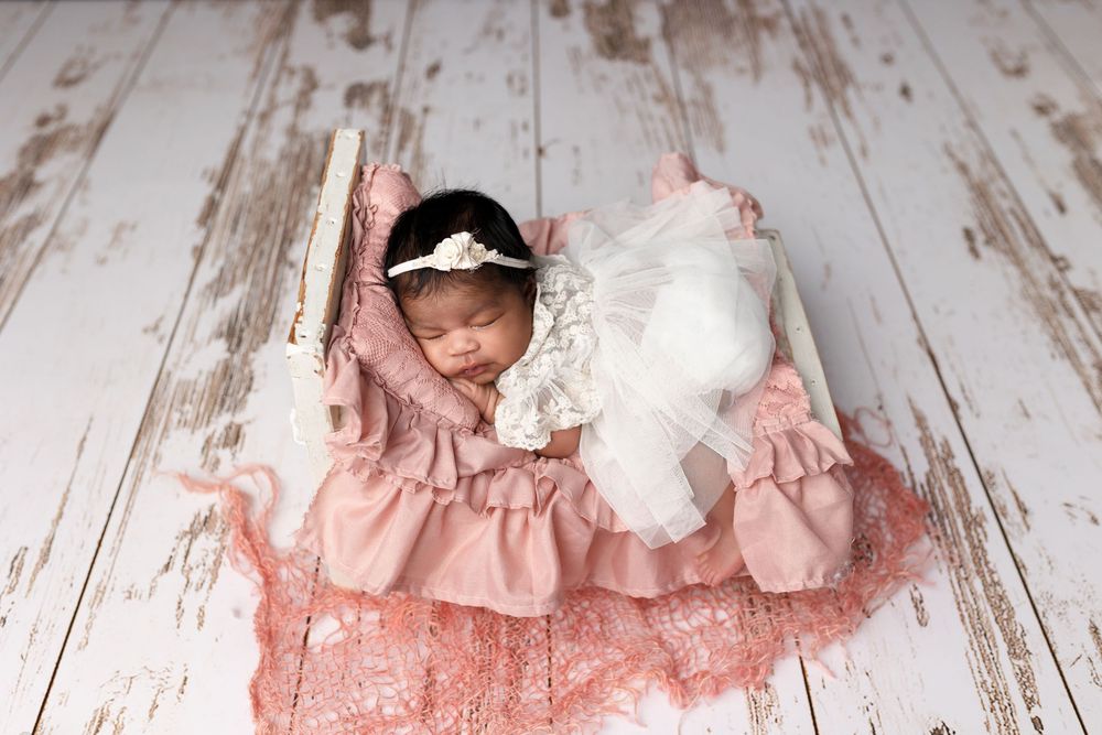A baby in a pink dress sits on a rustic wooden floor with white plush toys and floral decorations.