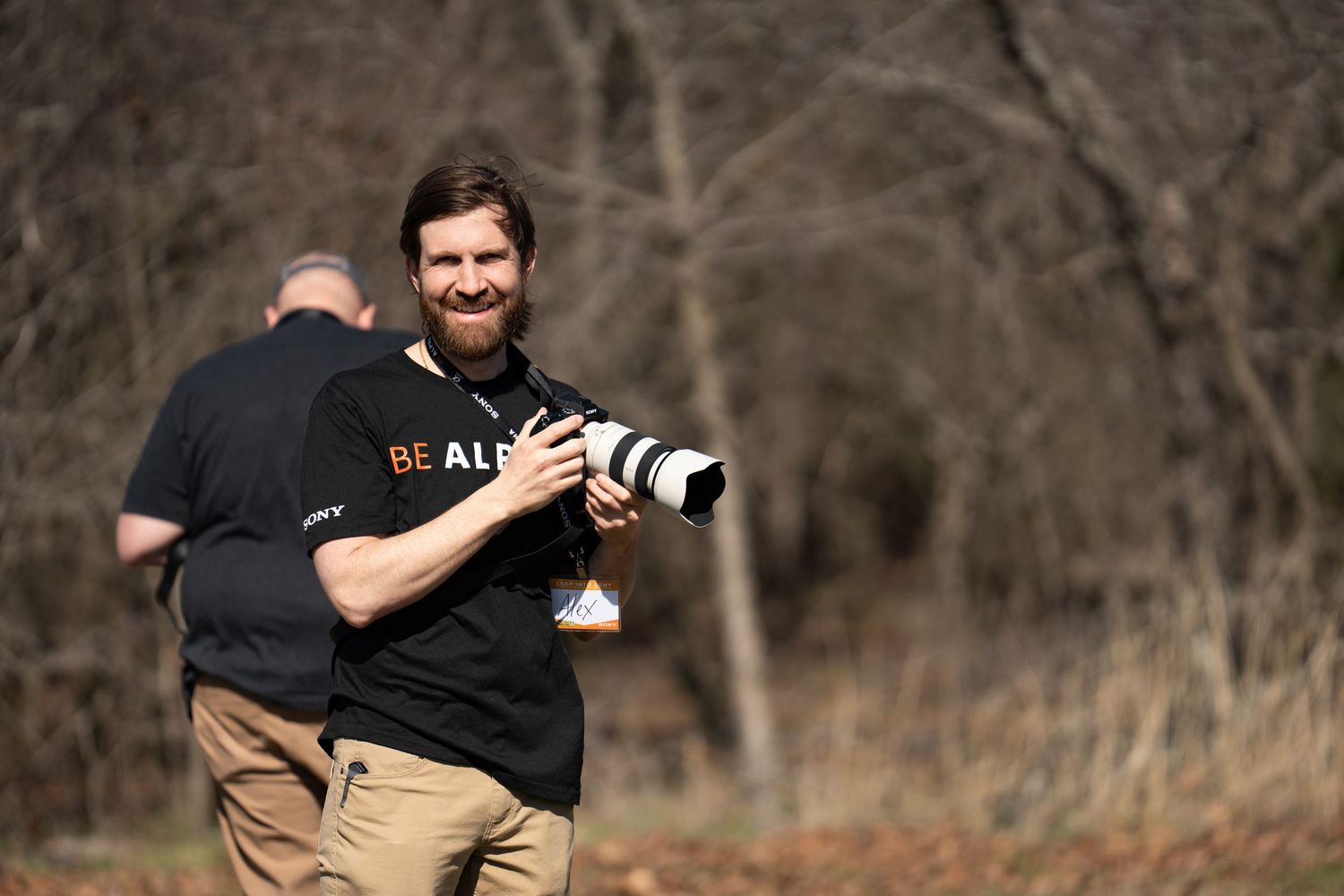 Arkansas nature photographer, Alex Kent grins at the camera holding a Sony camera with telephoto lens in bright sun