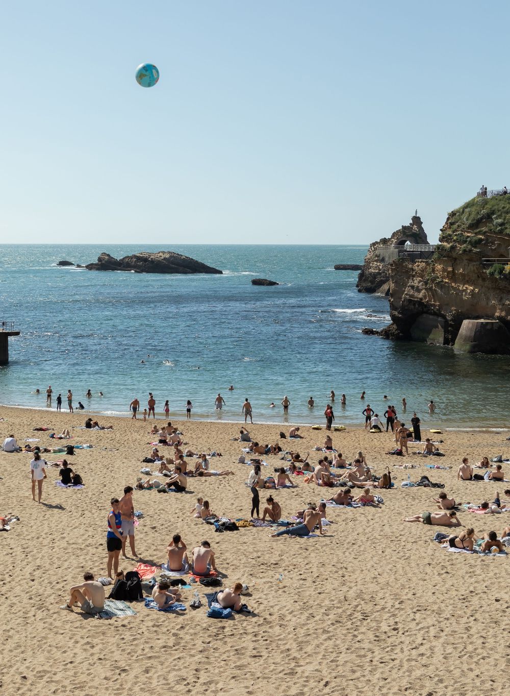 Beach goers relax and swim at a scenic rocky coastal beach with cliffs and blue waters under clear sunny skies.
