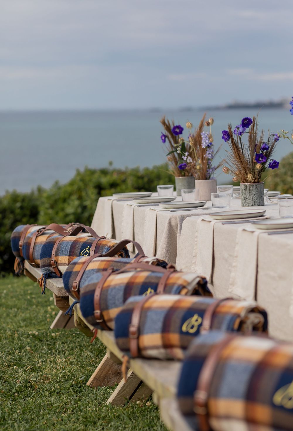 Rustic outdoor dining table settings with plaid blankets and floral centerpieces along the oceanfront grass lawn.