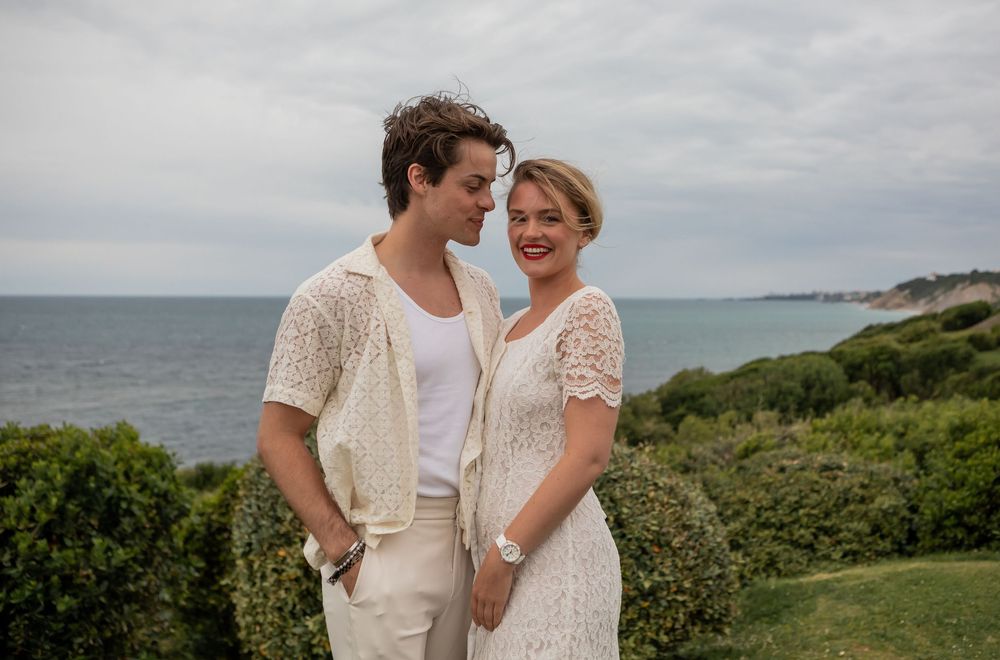 Two people in white attire share a celebratory toast outdoors by the ocean during their wedding ceremony.