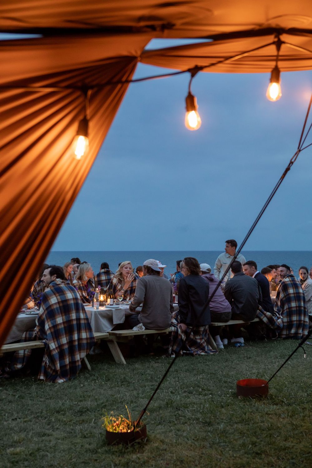 Outdoor dining setup at dusk with tents and string lights casting a warm glow over guests seated on the grass by the ocean.