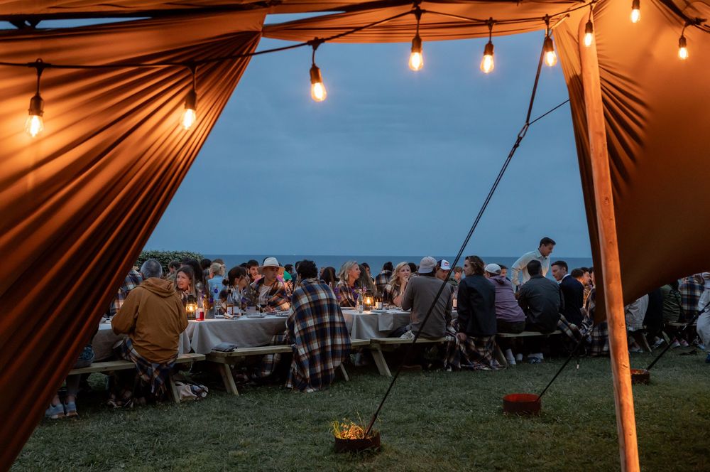 String lights illuminate an outdoor evening event under a large orange tent as guests gather at tables on the grass.