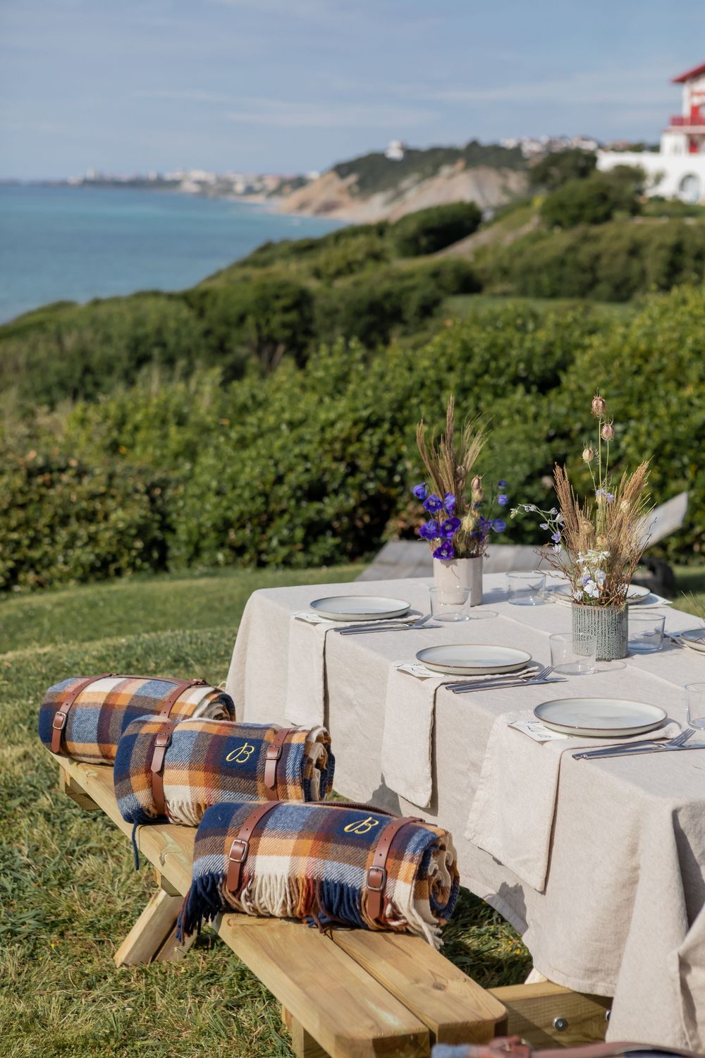Rustic outdoor table setting with plaid blanket and place settings against a coastal landscape background.