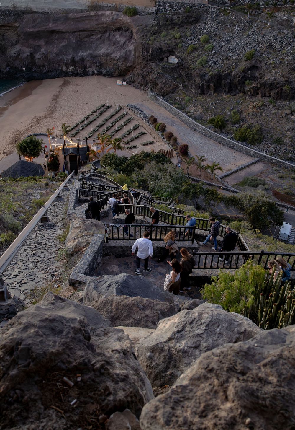 Aerial view of tourists on rocky terraced viewpoint overlooking a sandy valley with lush greenery in a coastal landscape.