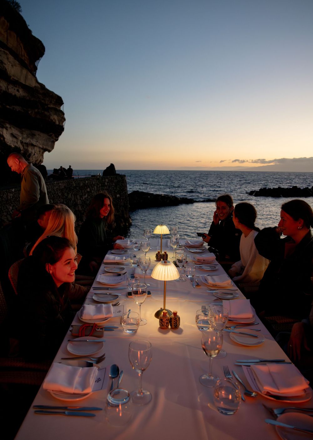 Elegant outdoor dinner table set by rocky ocean cliffs at dusk, lit by a glowing lamp with guests dining seaside.