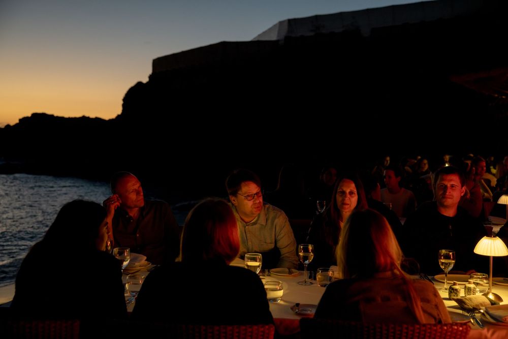 Group of people dining outdoors at dusk near rocky cliffs by the sea, illuminated by soft candlelight.