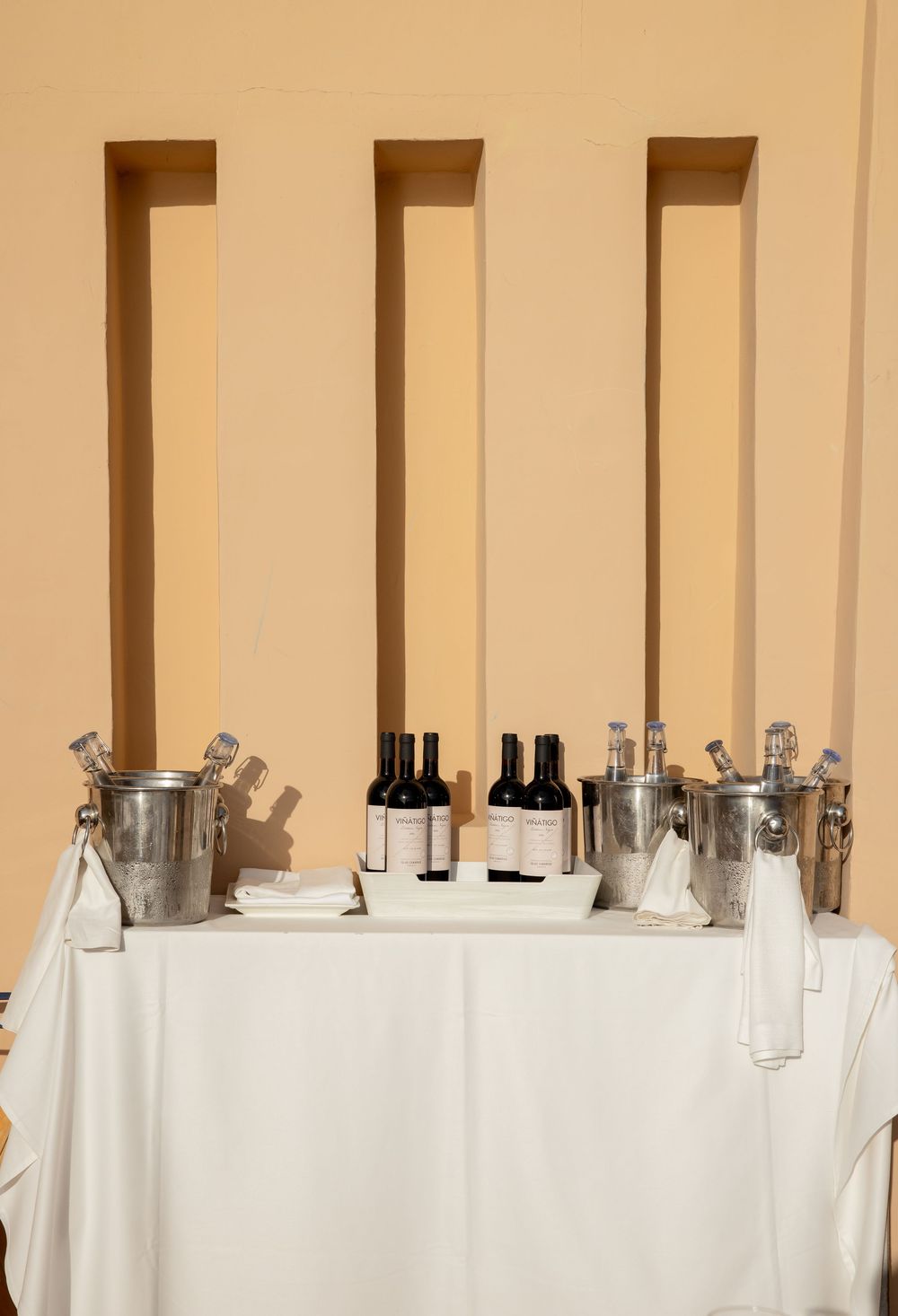 Elegant bar setup with wine bottles and silver ice buckets on a white linen table against a warm beige wall.