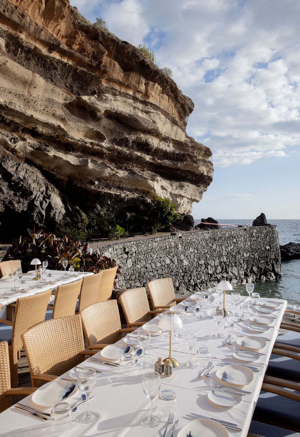 Elegant outdoor dining table set with white linens beside dramatic ocean cliffs and sea views under a partly cloudy sky.