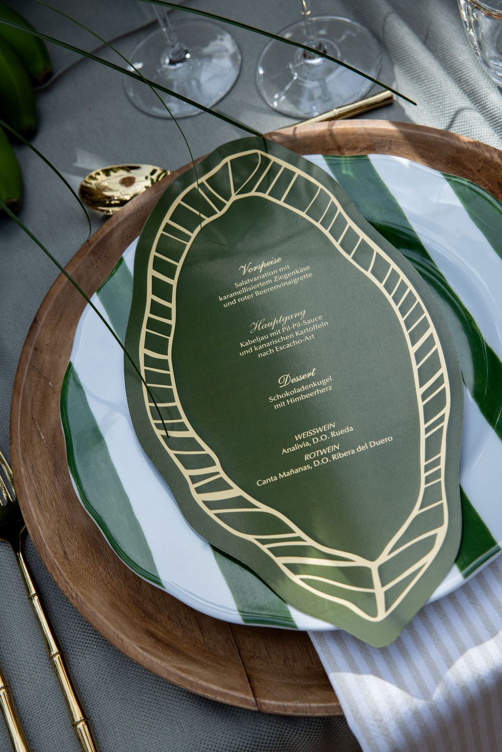 Elegant green and gold leaf-shaped wedding menu card on wooden charger plate with striped napkin and gold cutlery.