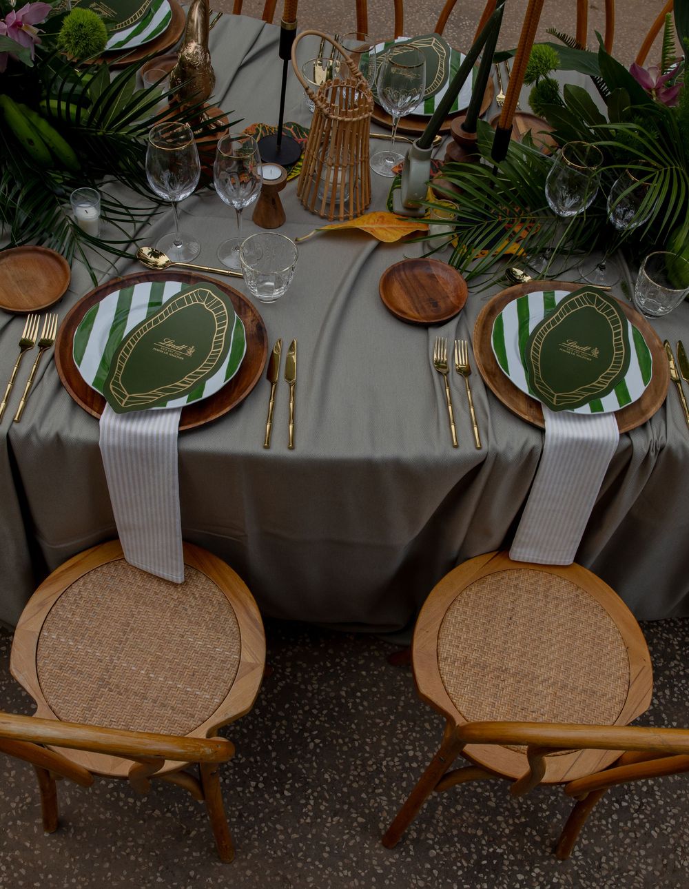 Elegant tropical table setting with green and white plates, gold cutlery, rattan chairs, and lush greenery centerpiece.