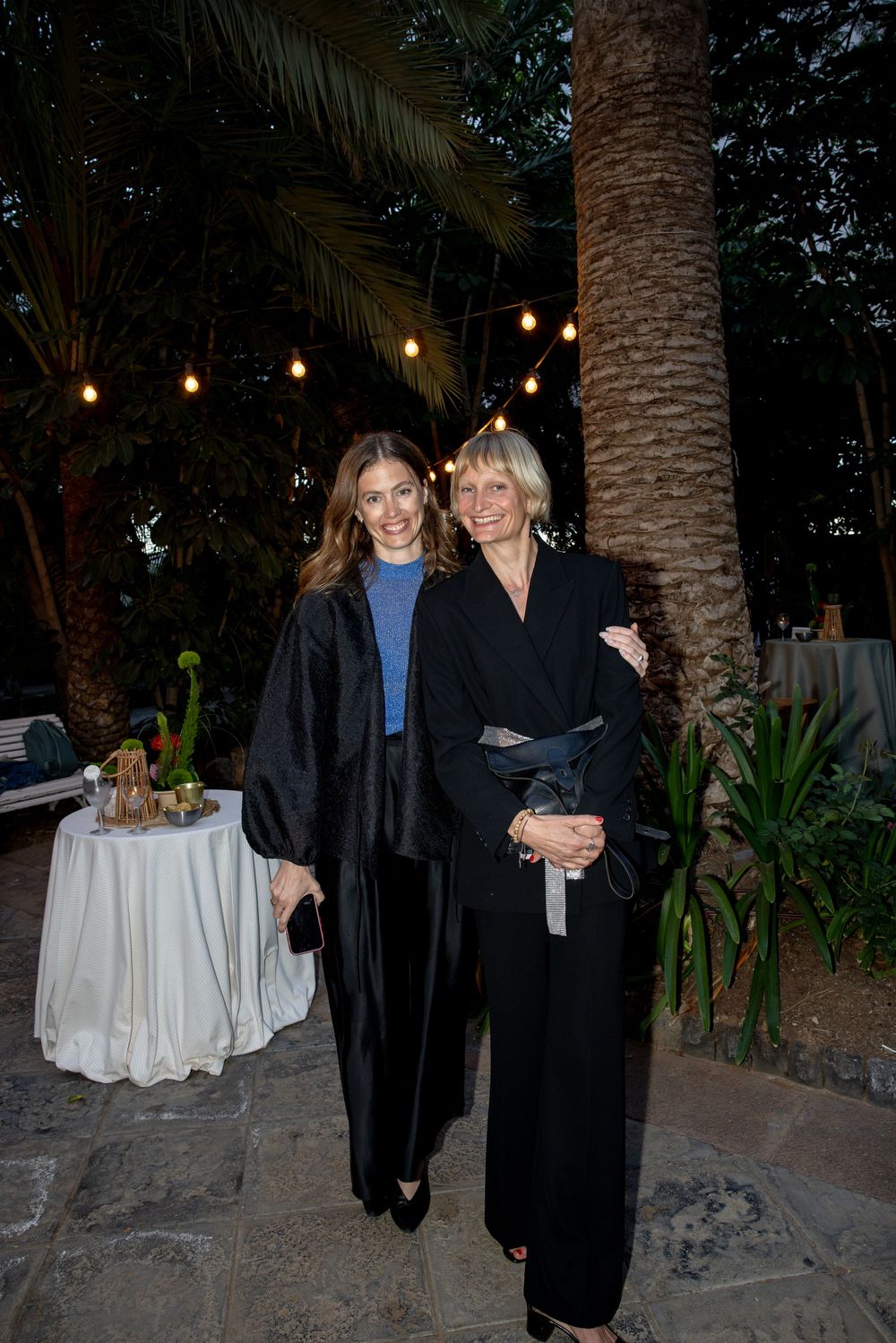 Two women in black outfits smiling outdoors at night under string lights surrounded by palm trees.