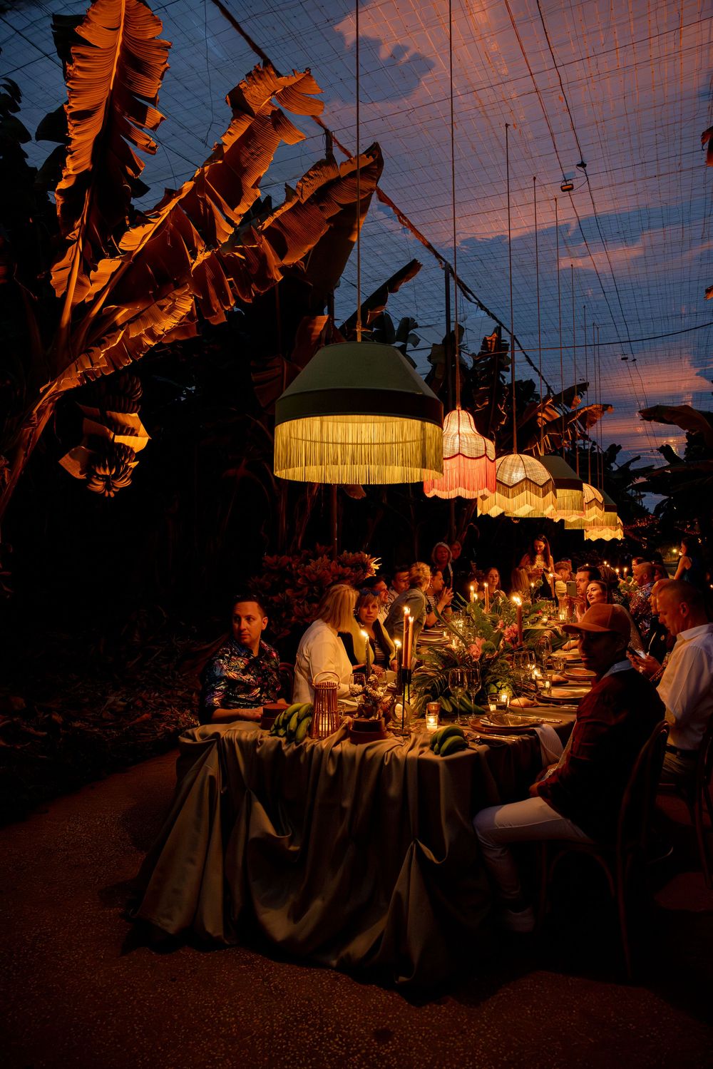 Elegant outdoor dinner party at dusk with hanging colorful lamps illuminating guests at a long decorated table.