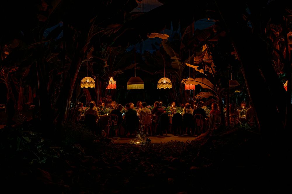 Dark atmospheric dining scene with guests seated at a long table under colorful stained glass windows in a tropical setting.