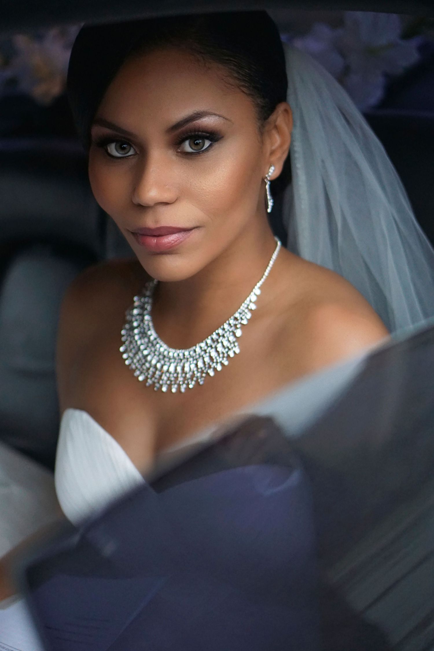 Stunning Black bride with diamond necklace and wedding veil posing in a car before her ceremony.