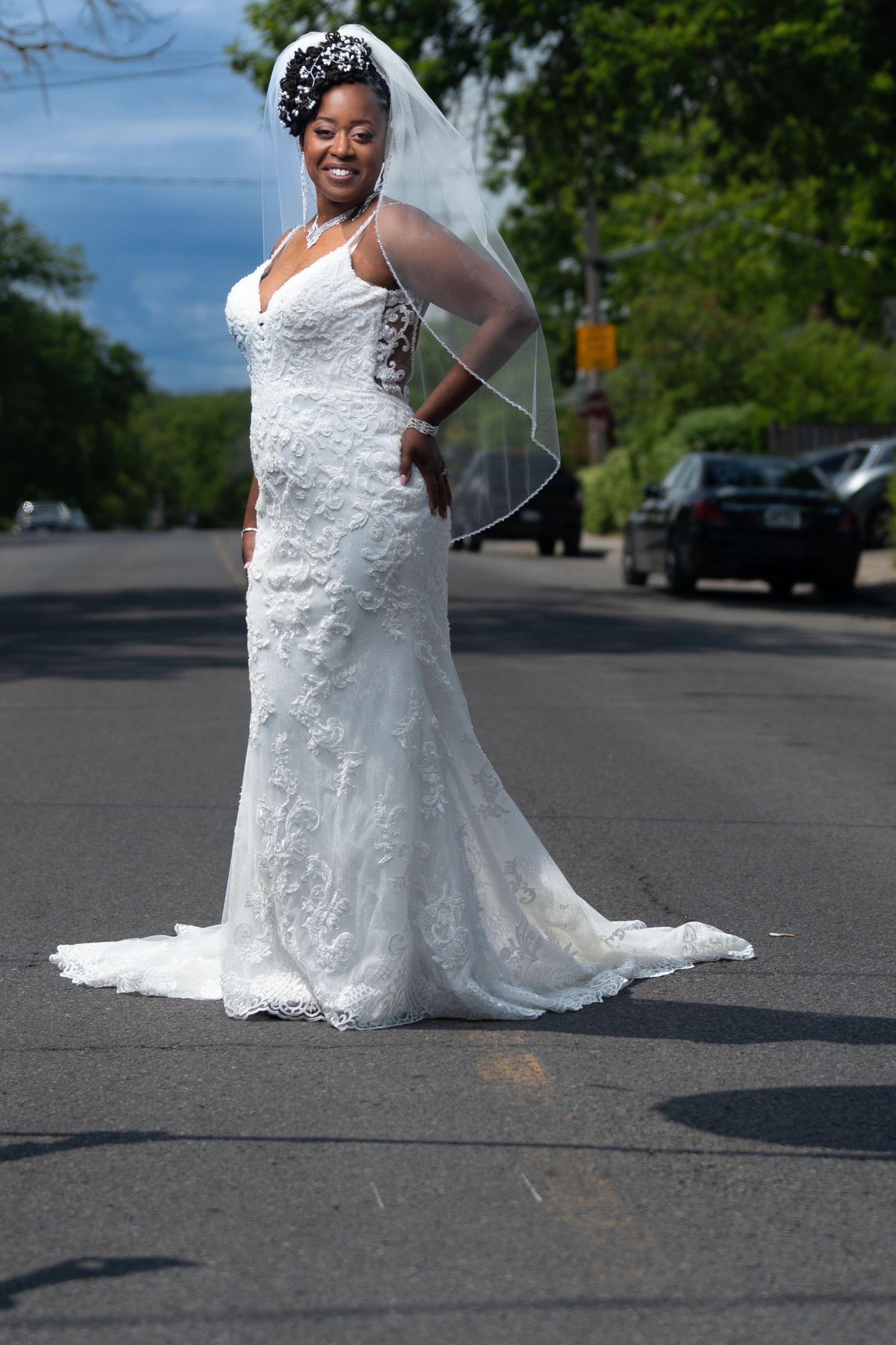 Beautiful Black bride in a white lace mermaid wedding dress and veil standing on a street.