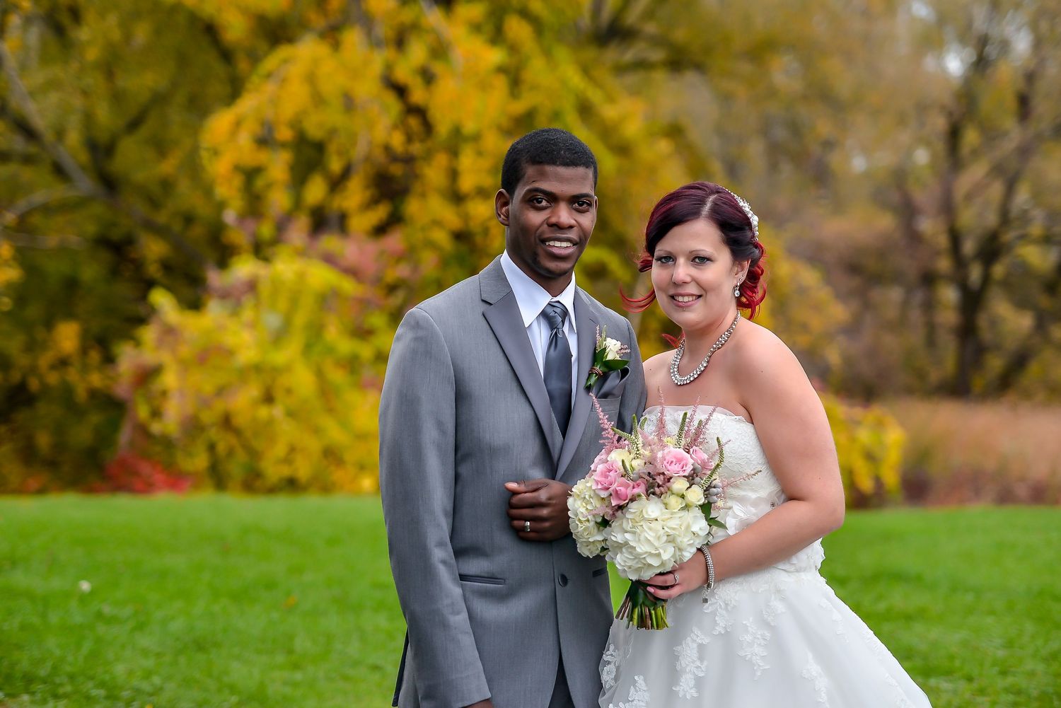 Interracial couple posing outdoors on wedding day, groom in gray suit, bride in white gown holding pink bouquet.