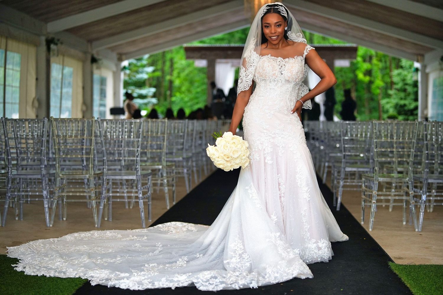 Bride in off-shoulder lace wedding dress with cathedral train poses at elegant outdoor ceremony.