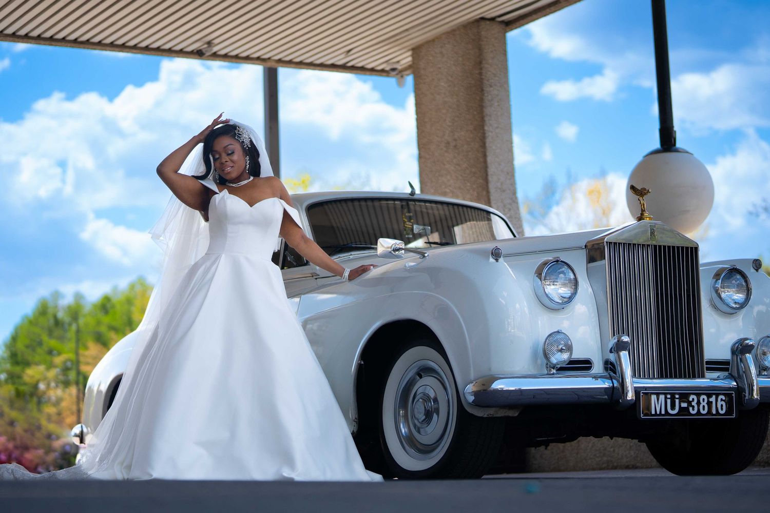 Bride in white wedding gown posing beside a classic white Rolls-Royce under a canopy on a sunny day.