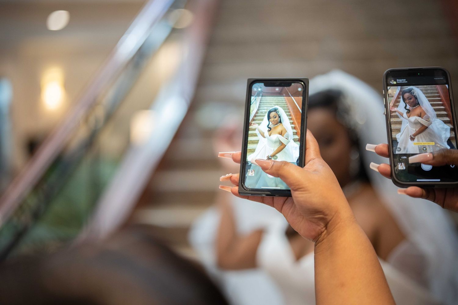 Hands holding smartphones photograph a bride in a white veil on a staircase.