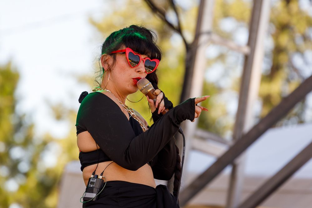 A performer in red sunglasses and black outfit sings on stage during an outdoor music festival.