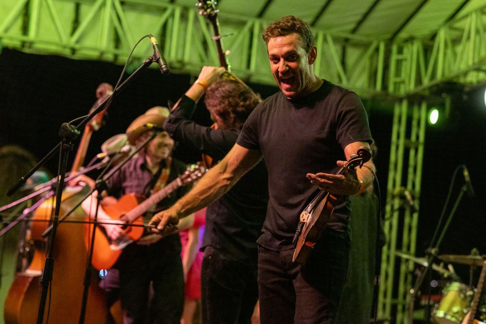 Musicians celebrate on stage during outdoor festival performance with green lighting.