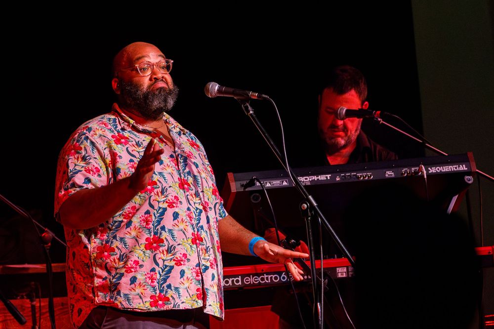 Musicians perform on stage with keyboard and microphone under red lighting during a live concert performance.