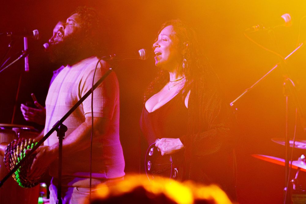 Stage performers in silhouette against vivid orange lighting during a dynamic live music performance.