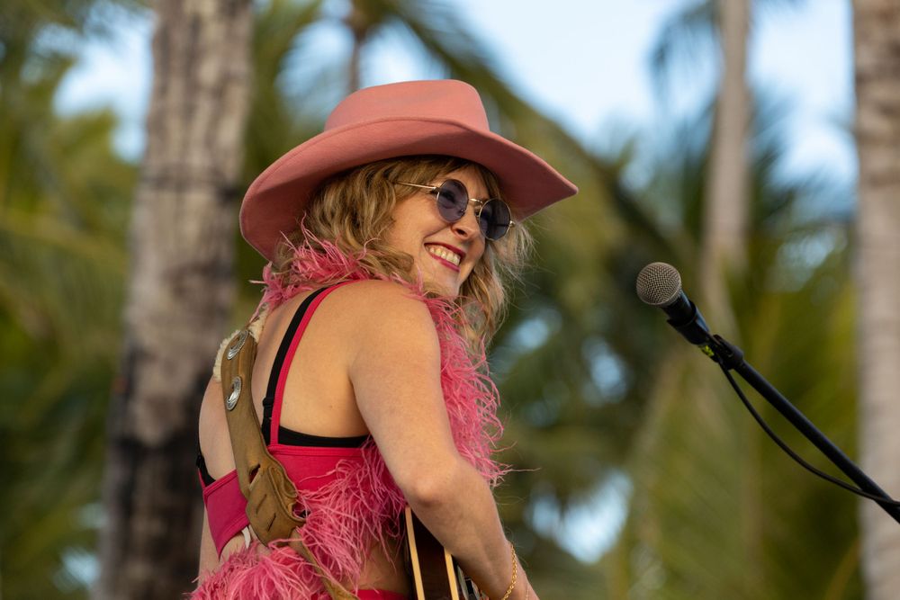 Musician in pink dress and cowboy hat performs outdoors with palm trees in background.