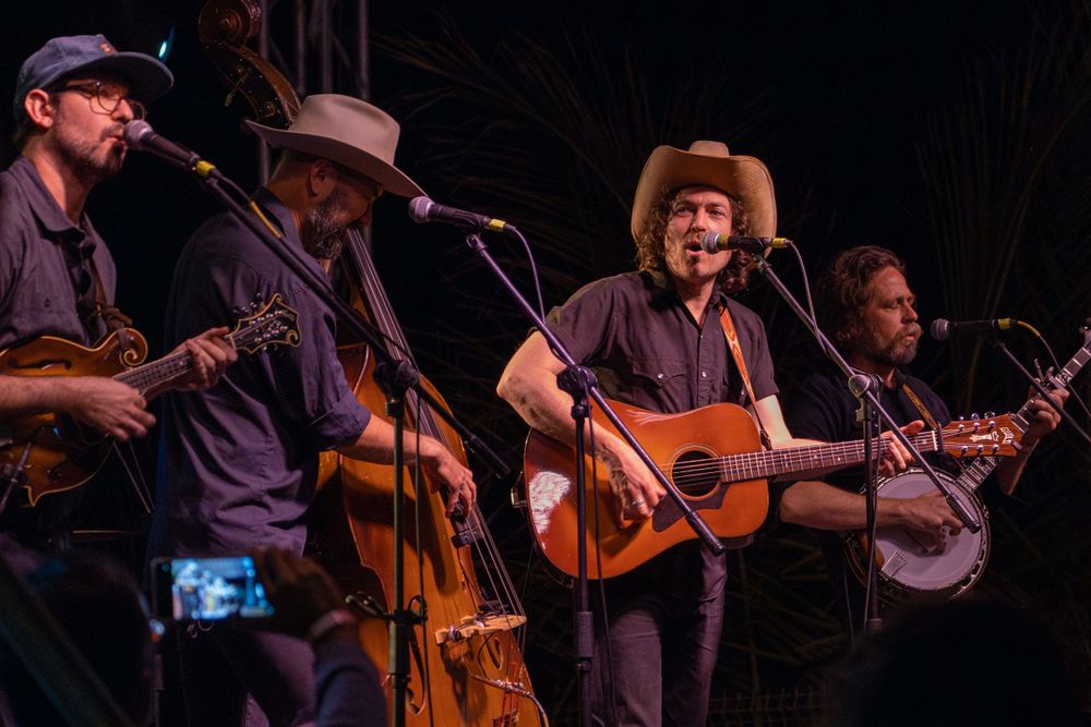 Folk band performs with acoustic instruments including banjo and upright bass on dark stage.