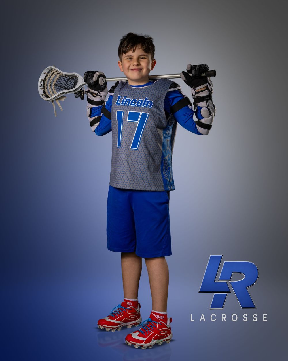 Young boy in Lincoln #17 lacrosse uniform holds stick behind shoulders, wearing blue shorts and red sneakers.