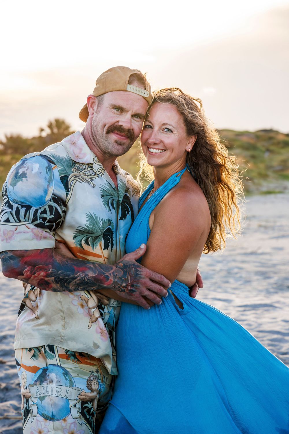 A couple embraces on a sandy beach at sunset wearing a blue dress and tropical print shirt.