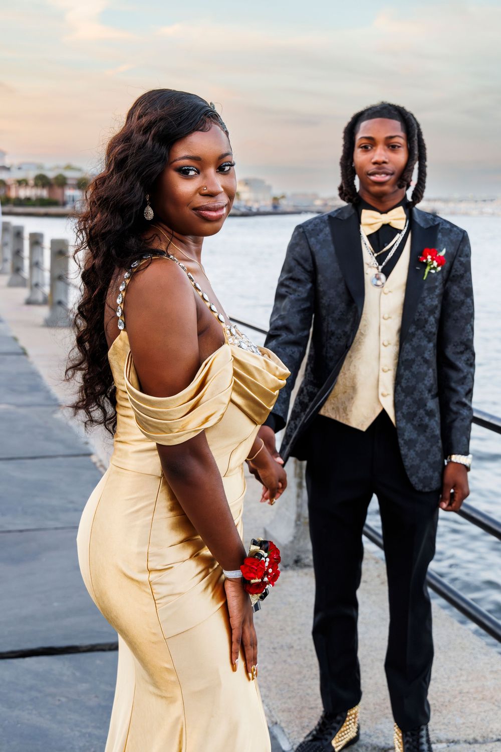 A couple poses together on a pier at sunset, wearing formal attire with a yellow dress and black tuxedo.