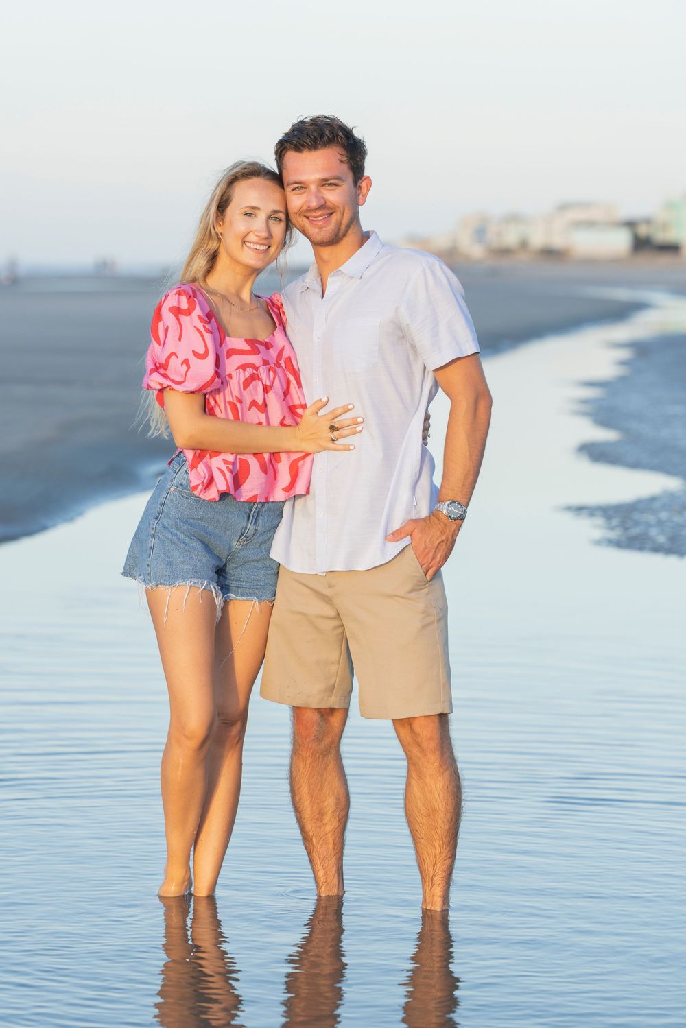 A couple stands together in shallow ocean water at the beach during sunset, wearing casual summer attire.