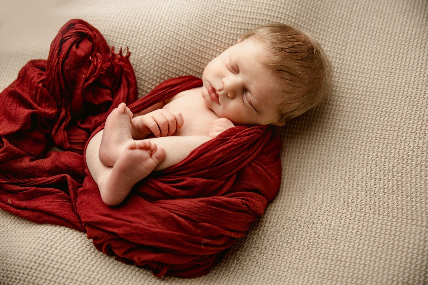 Peaceful sleeping baby wrapped in soft red velvet blanket on beige backdrop.